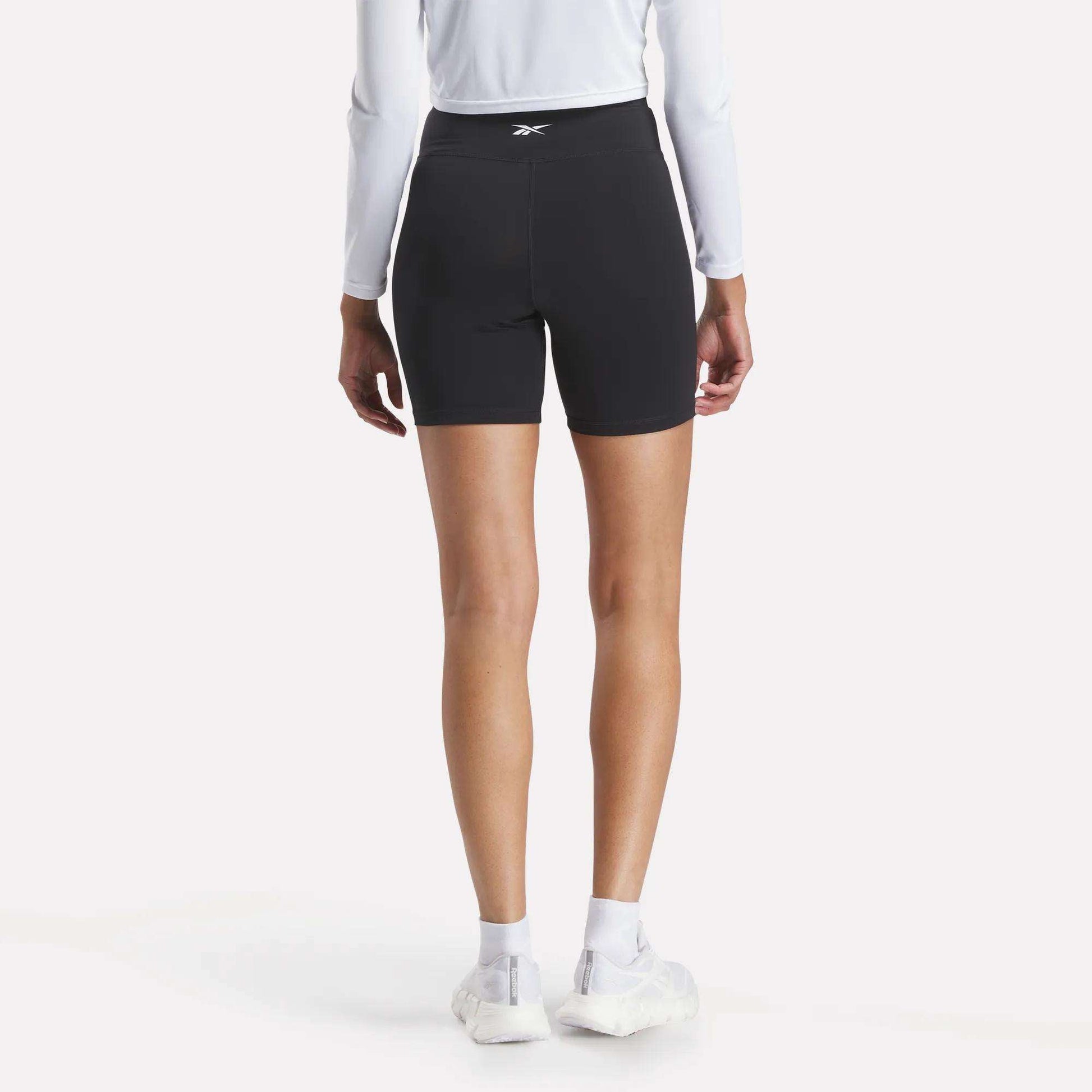 A person in Reebok Women's Core High-Rise Cycle Shorts, a long-sleeve white shirt, white socks, and white sneakers stands with their back to the camera against a plain light background.