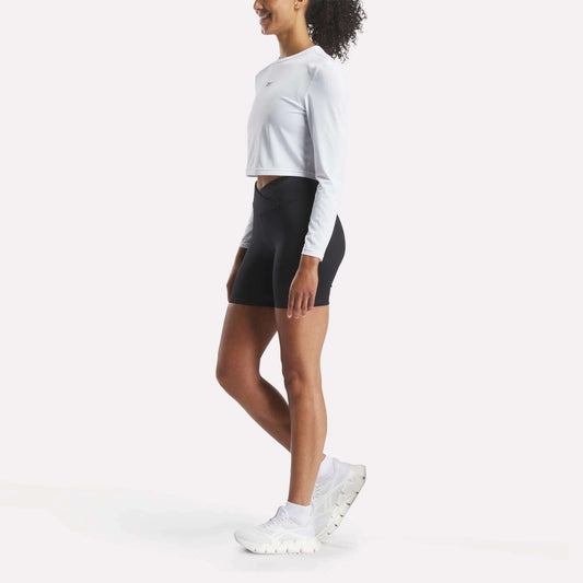 A woman stands sideways against a plain white background, wearing a long-sleeve white crop top, Reebok Women's Core High-Rise Cycle Shorts, and white athletic shoes.