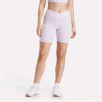A woman models Reebok Women's Core High-Rise Cycle Shorts in light purple with a crossover waistband, paired with a matching crop top, white ankle socks, and sneakers against a plain white background.