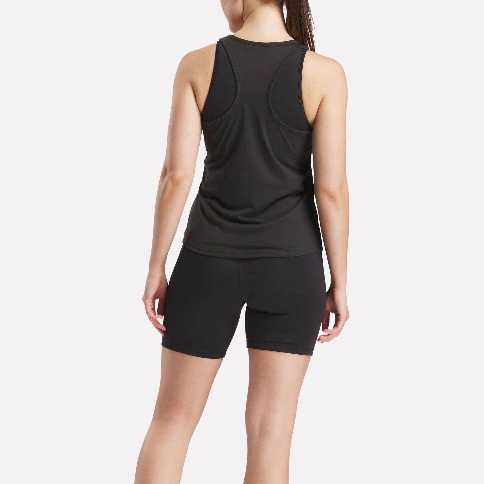 A woman faces away, wearing a Reebok Women's Core Tank Top and black sweat-wicking shorts against a plain light background.