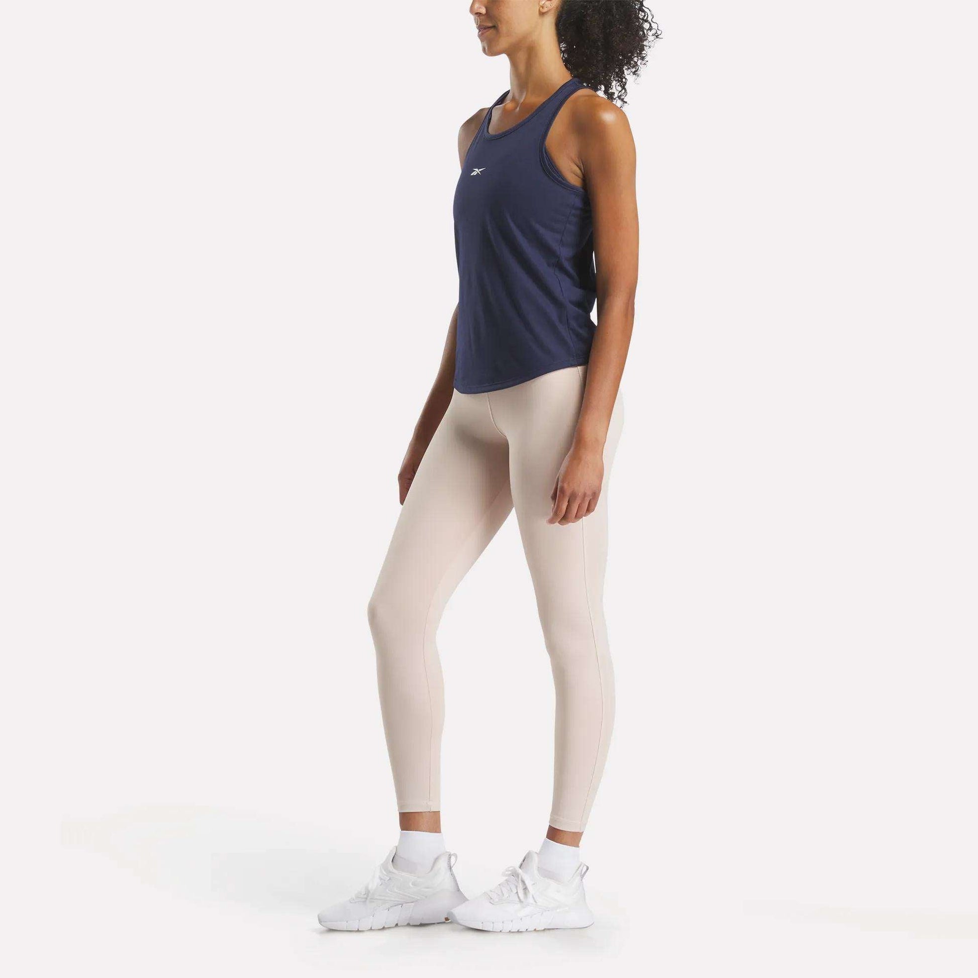 A woman poses in slim-fit athletic wear, wearing Reebok's Women's Core Tank Top in navy with sweat-wicking fabric, paired with light beige leggings and white sneakers against a white background.