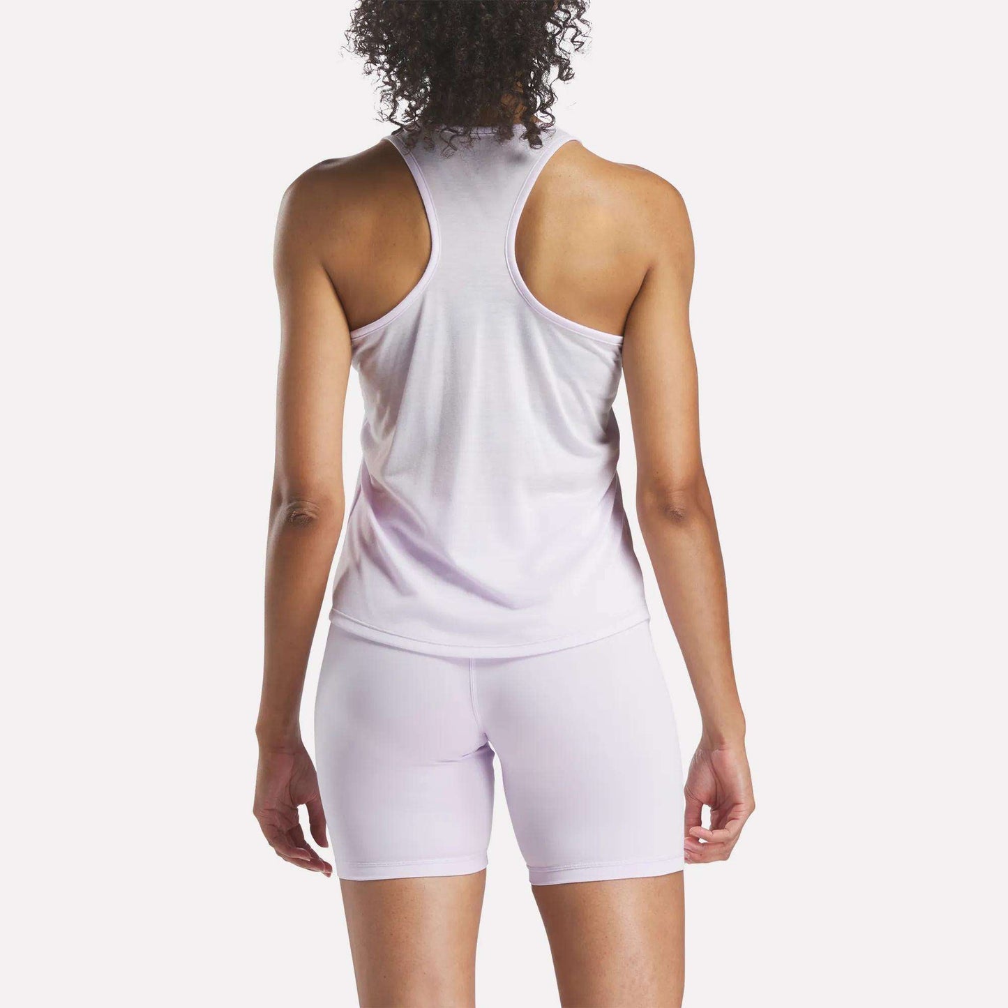 A person with curly hair is shown from the back, wearing the Reebok Women's Core Tank Top in light purple with matching fitted shorts, set against a plain white background.