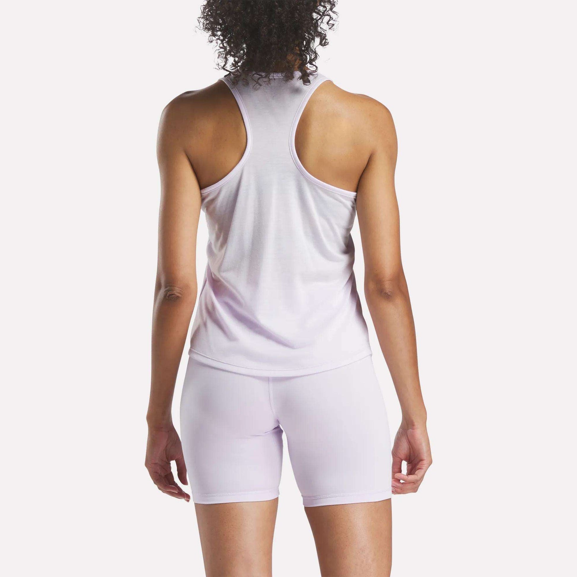 A person with curly hair is shown from the back, wearing the Reebok Women's Core Tank Top in light purple with matching fitted shorts, set against a plain white background.