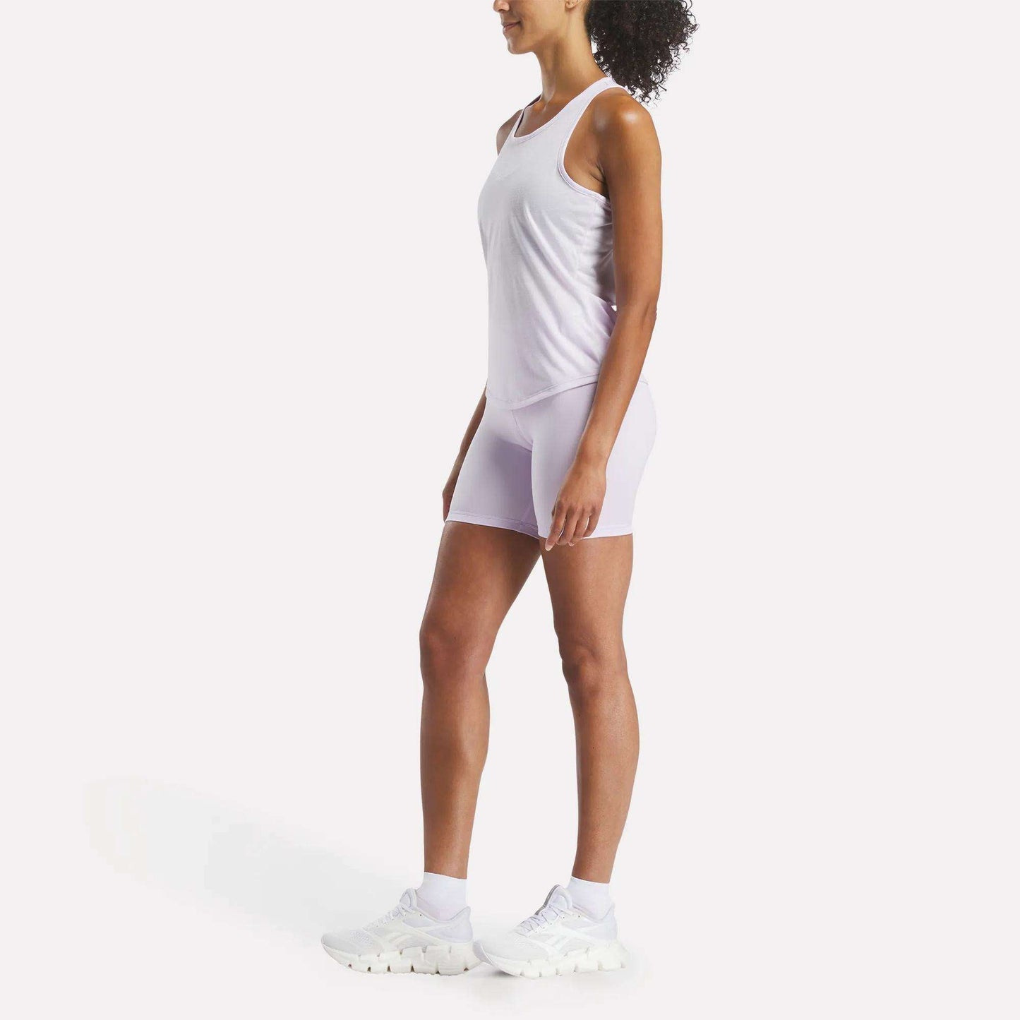 A woman stands in profile facing left, wearing a Reebok Women's Core Tank Top and fitted white shorts with white sneakers against a plain white background.