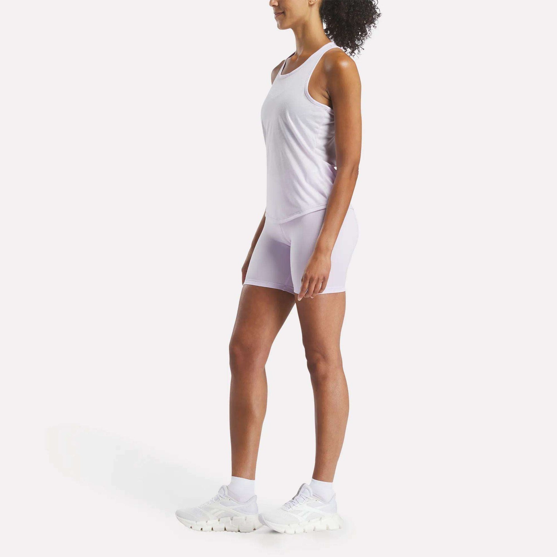 A woman stands in profile facing left, wearing a Reebok Women's Core Tank Top and fitted white shorts with white sneakers against a plain white background.