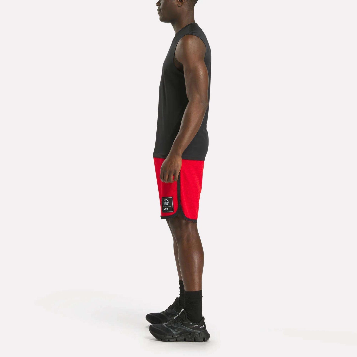 A man stands in profile facing left, wearing a black sleeveless shirt, Reebok Men's Basketball Mesh Shorts, black socks, and black sneakers against a plain white background.