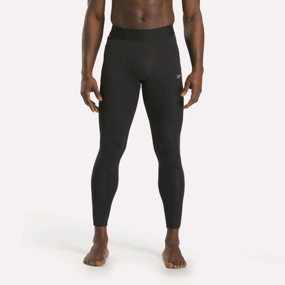 A person stands barefoot against a plain white background, wearing Reebok Men's Core Training Tights in black with a small white logo on the left thigh.