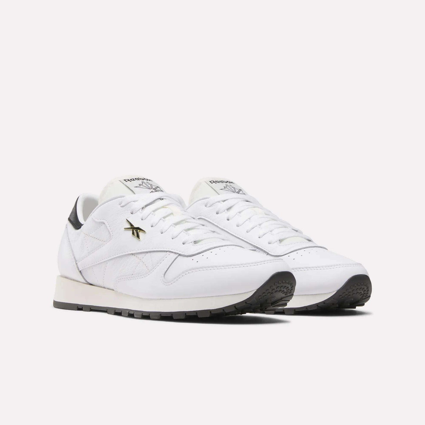 The Reebok Classic Leather Shoes showcase a retro running style with a white leather upper, black soles, gold logo accents, and white laces, set against a plain light background.