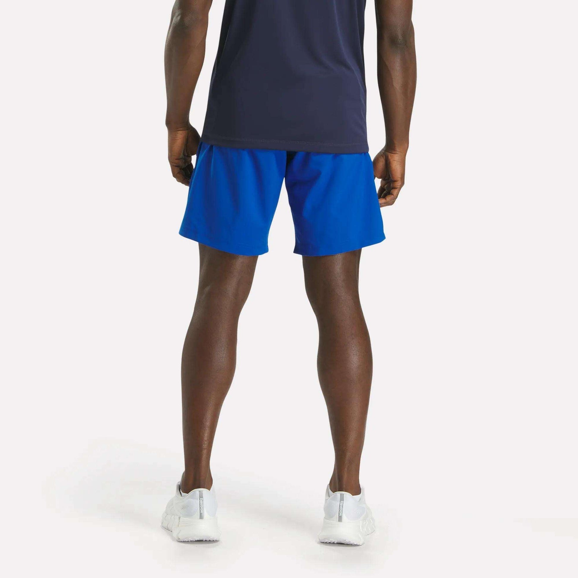 A man faces away, wearing a navy shirt and bright blue Reebok Men's Workout Ready Woven Shorts with white sneakers, set against a plain light background.