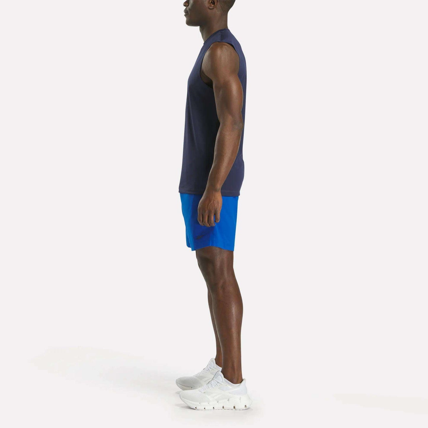 A man in profile facing left wears a sleeveless navy shirt, Reebok Men's Workout Ready Woven Shorts in bright blue, and white sneakers, standing against a plain white background.