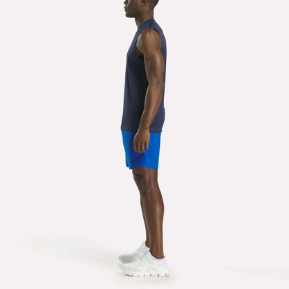 A man in profile facing left wears a sleeveless navy shirt, Reebok Men's Workout Ready Woven Shorts in bright blue, and white sneakers, standing against a plain white background.