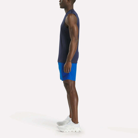 A man in profile facing left wears a sleeveless navy shirt, Reebok Men's Workout Ready Woven Shorts in bright blue, and white sneakers, standing against a plain white background.