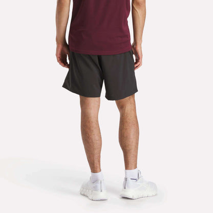 A person stands with their back to the camera, wearing a burgundy shirt and Reebok Men's Workout Ready Woven Shorts, known for sweat-wicking comfort, plus white socks and sneakers, against a plain light background.