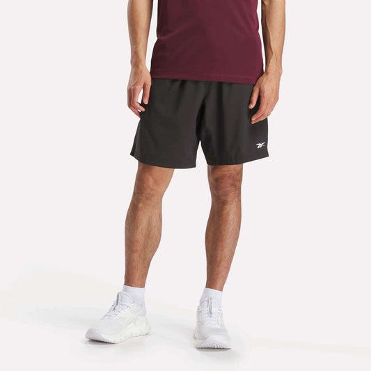 A person models the Reebok Men's Workout Ready Woven Shorts, shown from the waist down against a plain white background, paired with a burgundy T-shirt and white sneakers.