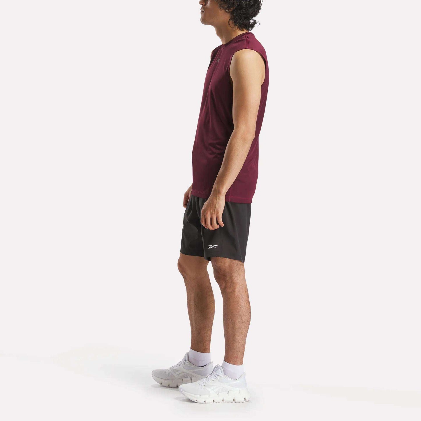 A man in profile wears a sleeveless burgundy athletic shirt, Reebok Men's Workout Ready Woven Shorts, white socks, and white athletic shoes against a plain white background.
