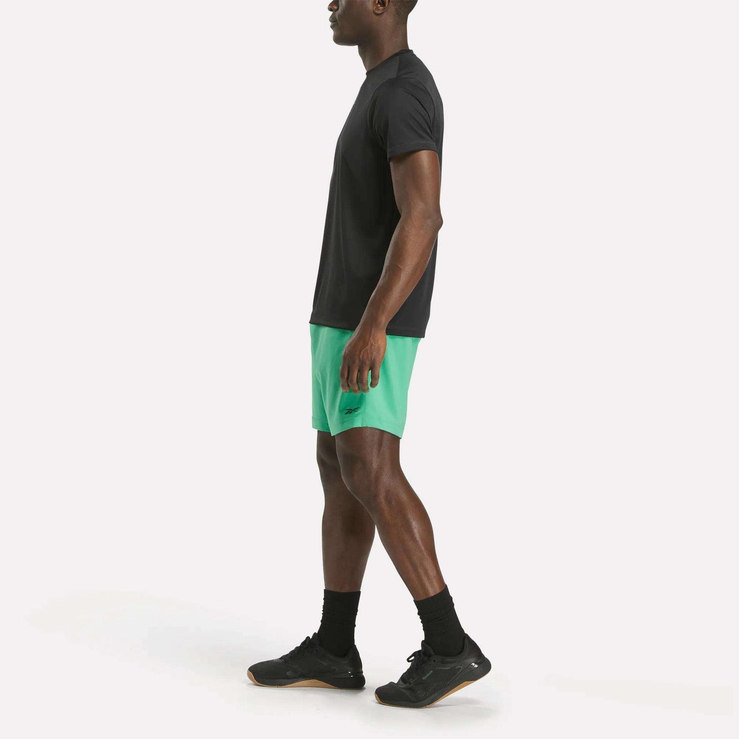 A man stands in profile wearing a black Reebok Men's ID Train Short Sleeve Tech Tee, green athletic shorts, black socks, and black sneakers against a plain white background.