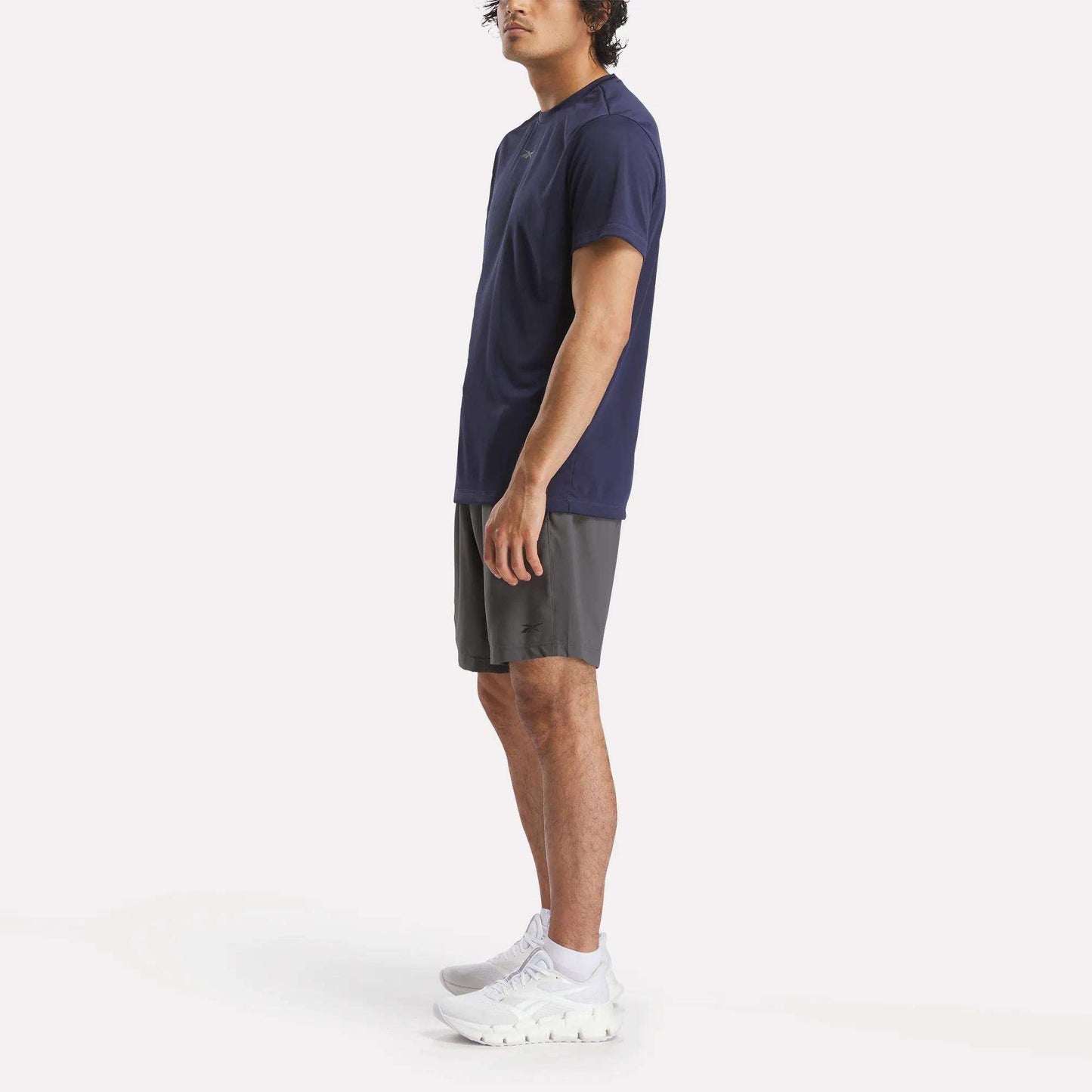 A man in profile wears a navy Reebok Men's ID Train Short Sleeve Tech Tee, gray shorts, and white athletic shoes against a plain white background.
