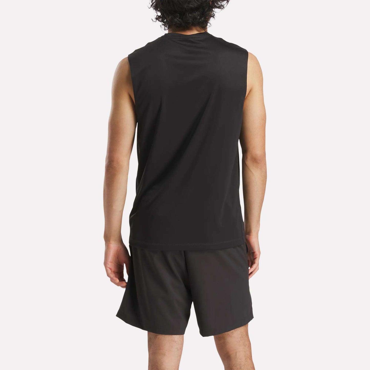 A person with curly hair is seen from the back, wearing a Reebok Men's Train Sleeveless Tech Tee and black lightweight polyester shorts, standing against a plain light background.