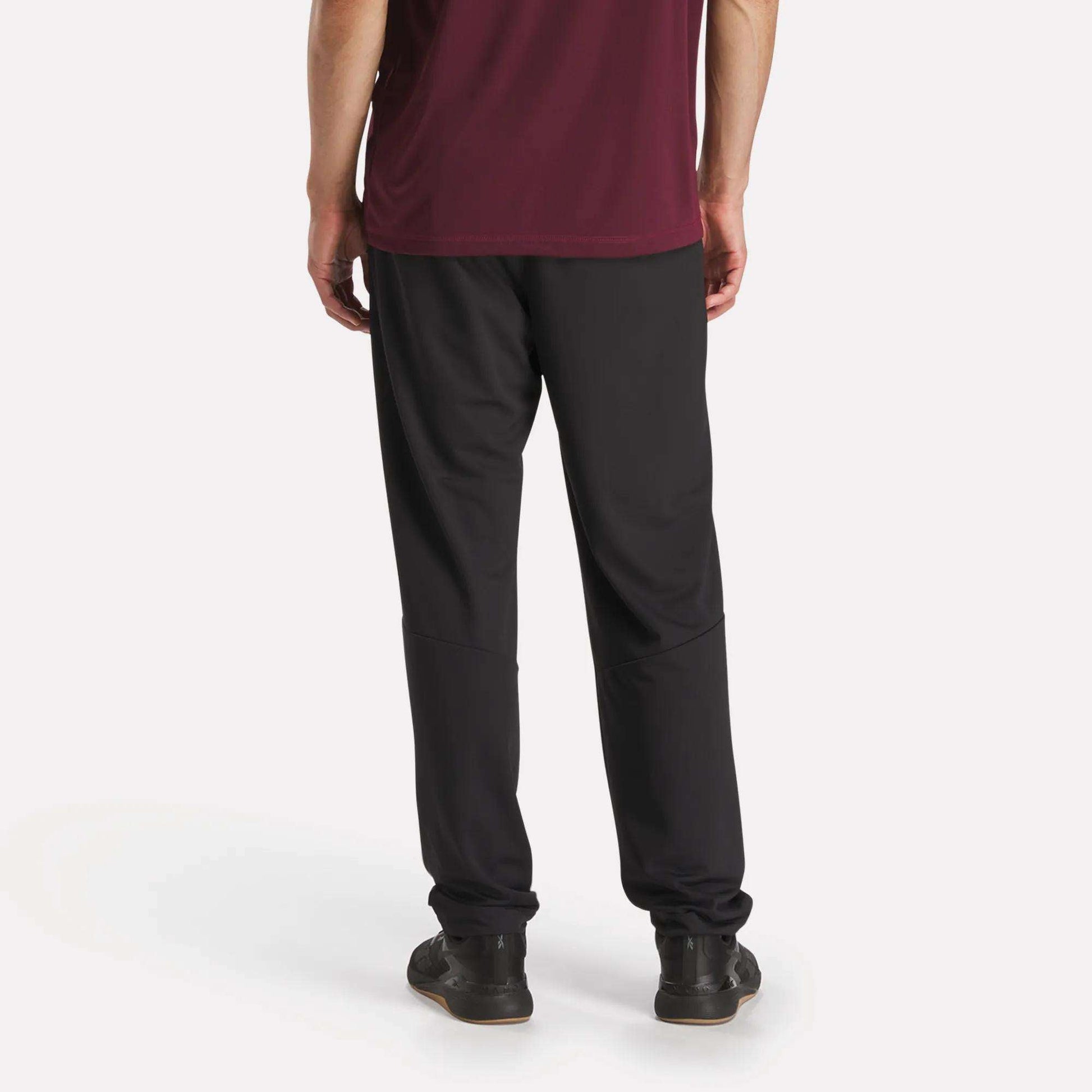 A person stands with their back to the camera, wearing a maroon shirt and black Reebok Men's Train Knit Pants with black sneakers, set against a plain white background.