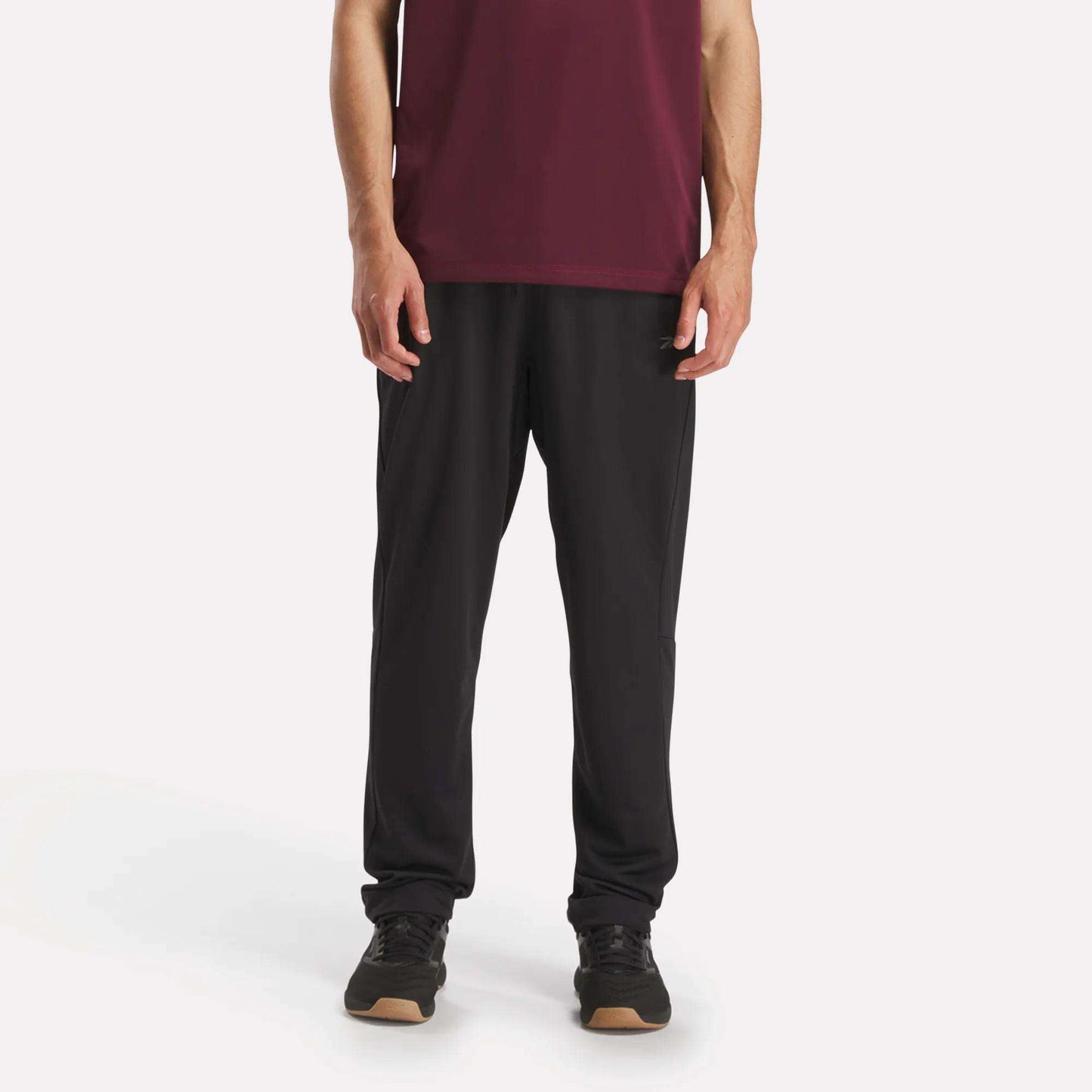 Against a plain white background, a person wears Reebok Men's Train Knit Pants with black sneakers and a maroon short-sleeve shirt. The photo, cropped at the shoulders, focuses on the lower half of the body.