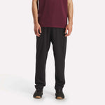 Against a plain white background, a person wears Reebok Men's Train Knit Pants with black sneakers and a maroon short-sleeve shirt. The photo, cropped at the shoulders, focuses on the lower half of the body.