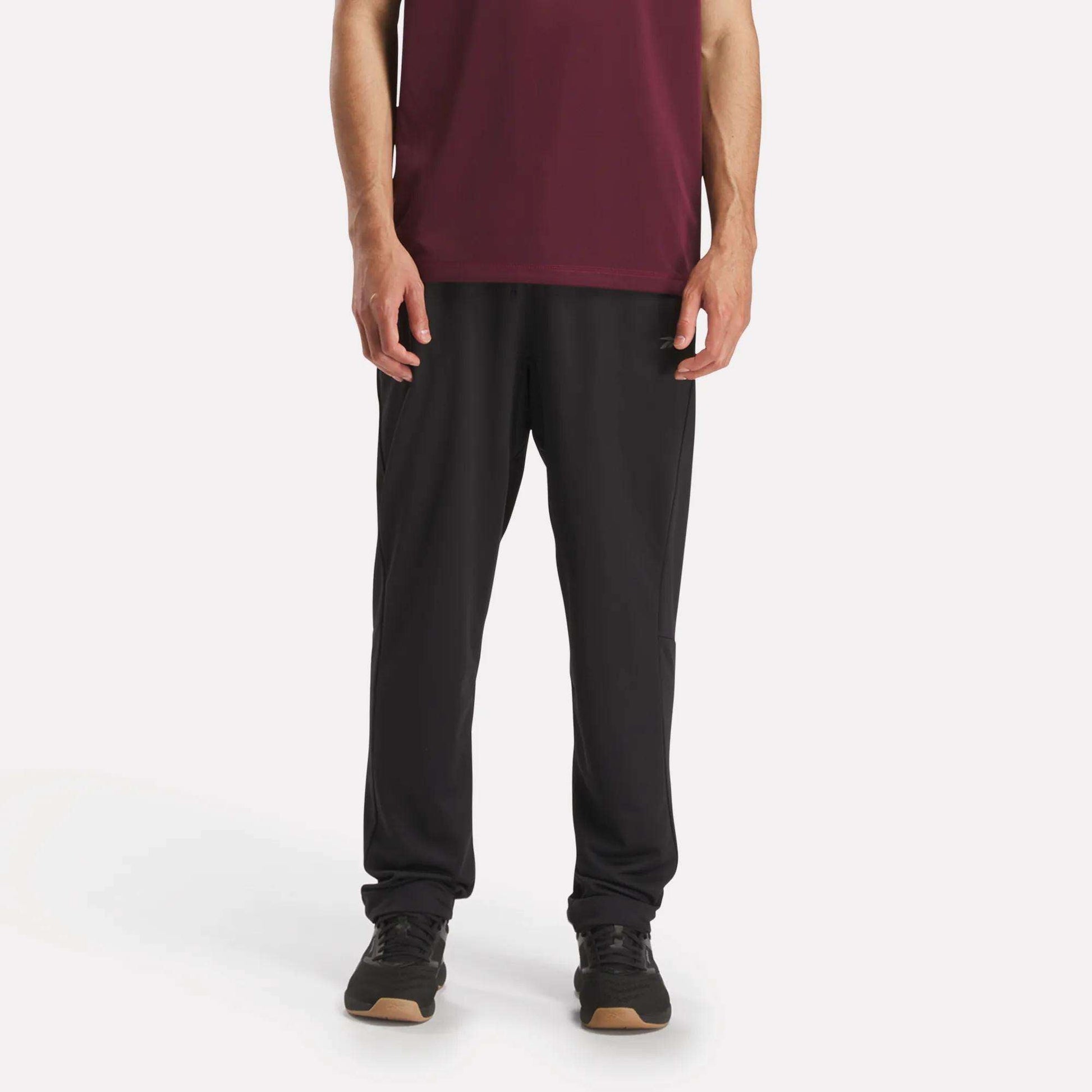 Against a plain white background, a person wears Reebok Men's Train Knit Pants with black sneakers and a maroon short-sleeve shirt. The photo, cropped at the shoulders, focuses on the lower half of the body.