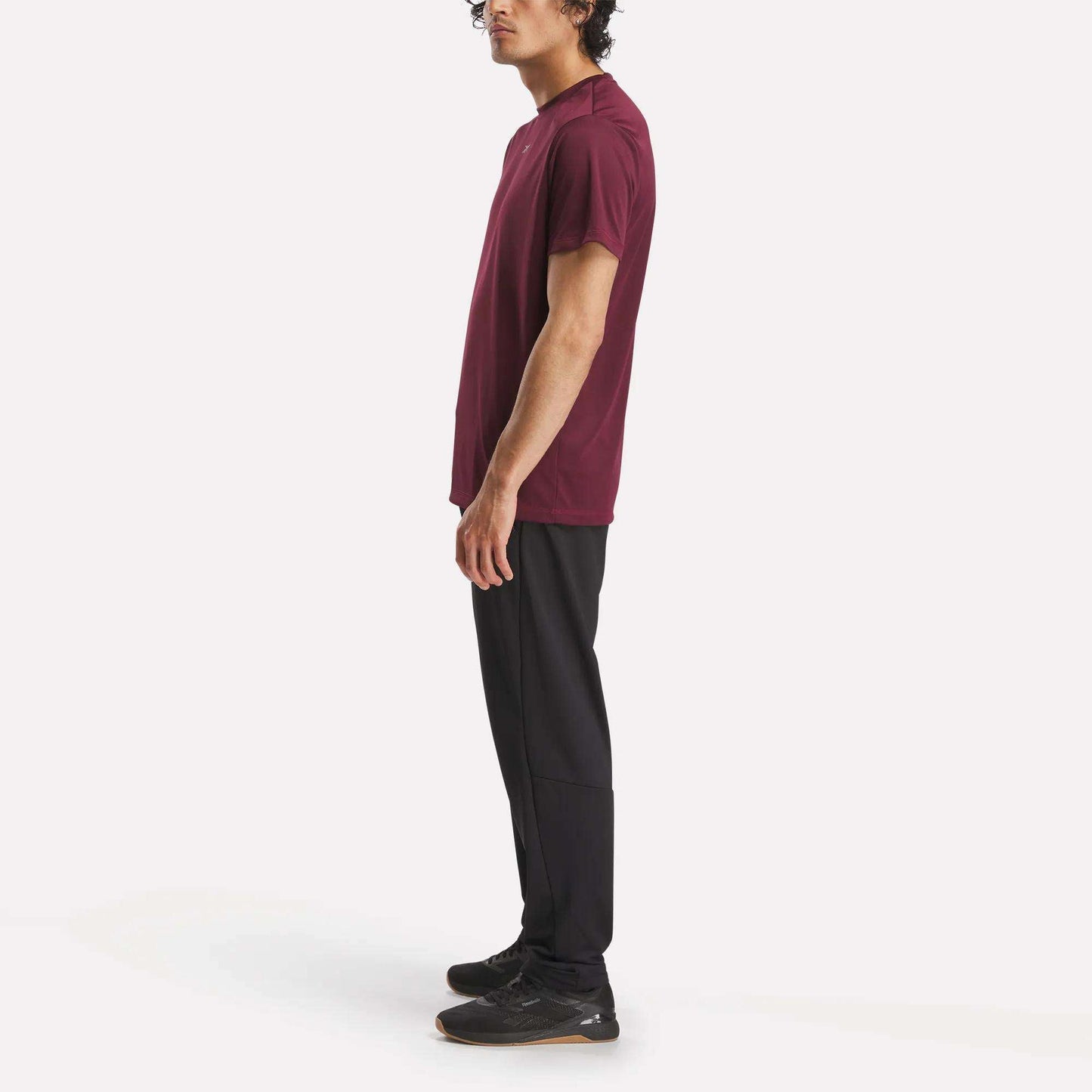 A man stands in profile wearing a burgundy t-shirt, black Reebok Men's Train Knit Pants, and black sneakers against a plain white background.