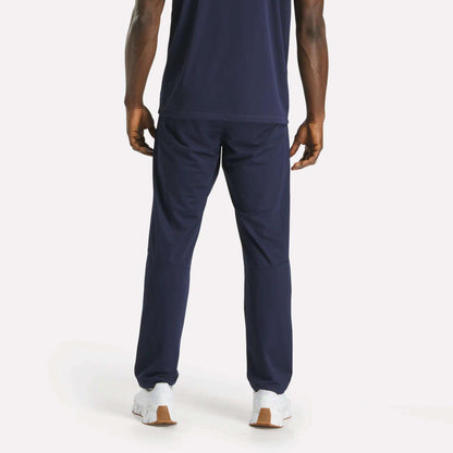 A man faces away, wearing a navy blue T-shirt and Reebok Men's Train Knit Pants with white sneakers on a plain light background.