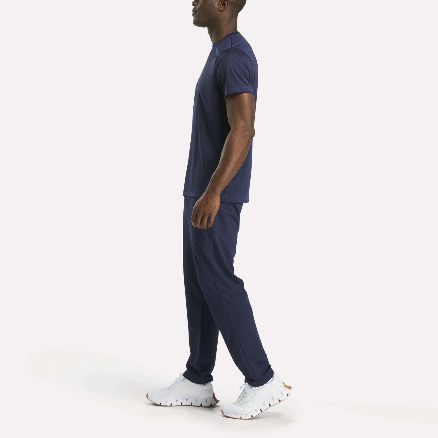 A man stands sideways wearing Reebok Men's Train Knit Pants in navy blue with a matching T-shirt and white athletic shoes, set against a plain white background.