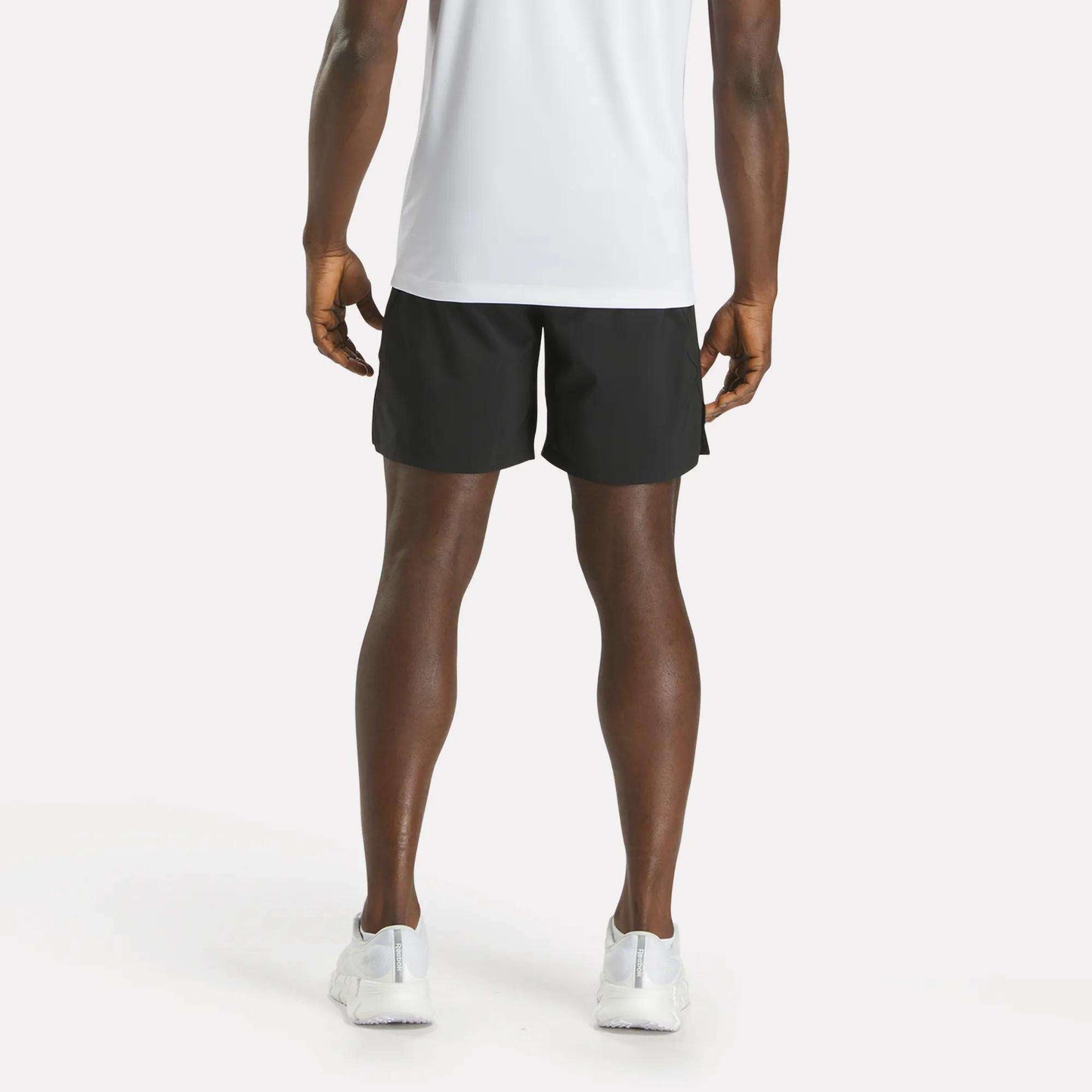 A person stands facing away from the camera, wearing a white T-shirt, Reebok Men's Athlete Speed Shorts in black, and white sneakers against a plain light background.