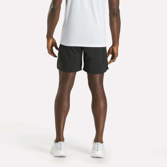 A person stands facing away from the camera, wearing a white T-shirt, Reebok Men's Athlete Speed Shorts in black, and white sneakers against a plain light background.