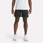 A person is shown from the shoulders down, facing forward against a plain white background, wearing a white shirt, white athletic shoes, and black Reebok Men's Athlete Speed Shorts featuring RBK-DRY technology.