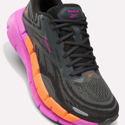 A close-up of the Reebok Women's FloatZig Double Running Shoe in black and gray with pink and orange accents, black laces, and visible Reebok logos, designed for max cushioning, set against a white background.