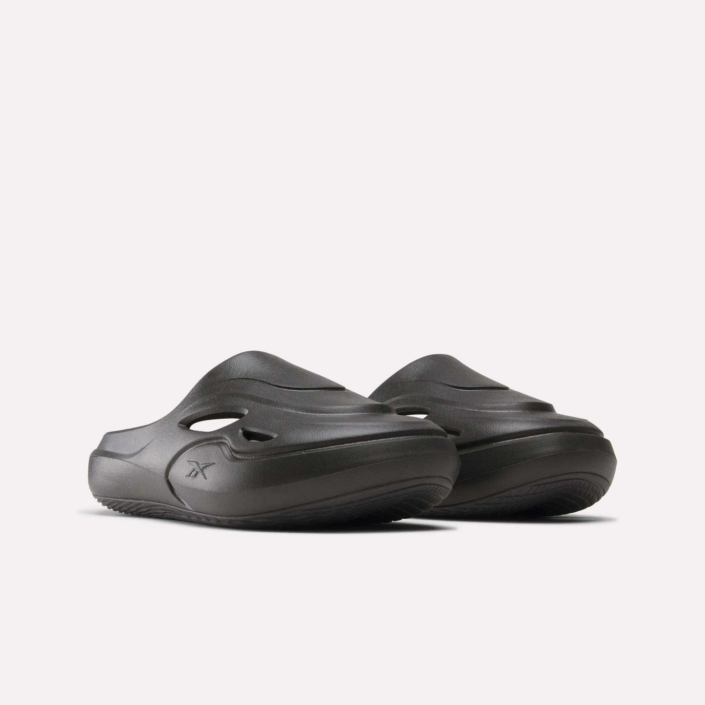 The Reebok DMX Recovery Mules are chunky dark gray molded EVA slip-on slides with side cutouts and grooved soles, shown against a plain light background.