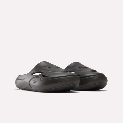 The Reebok DMX Recovery Mules are chunky dark gray molded EVA slip-on slides with side cutouts and grooved soles, shown against a plain light background.