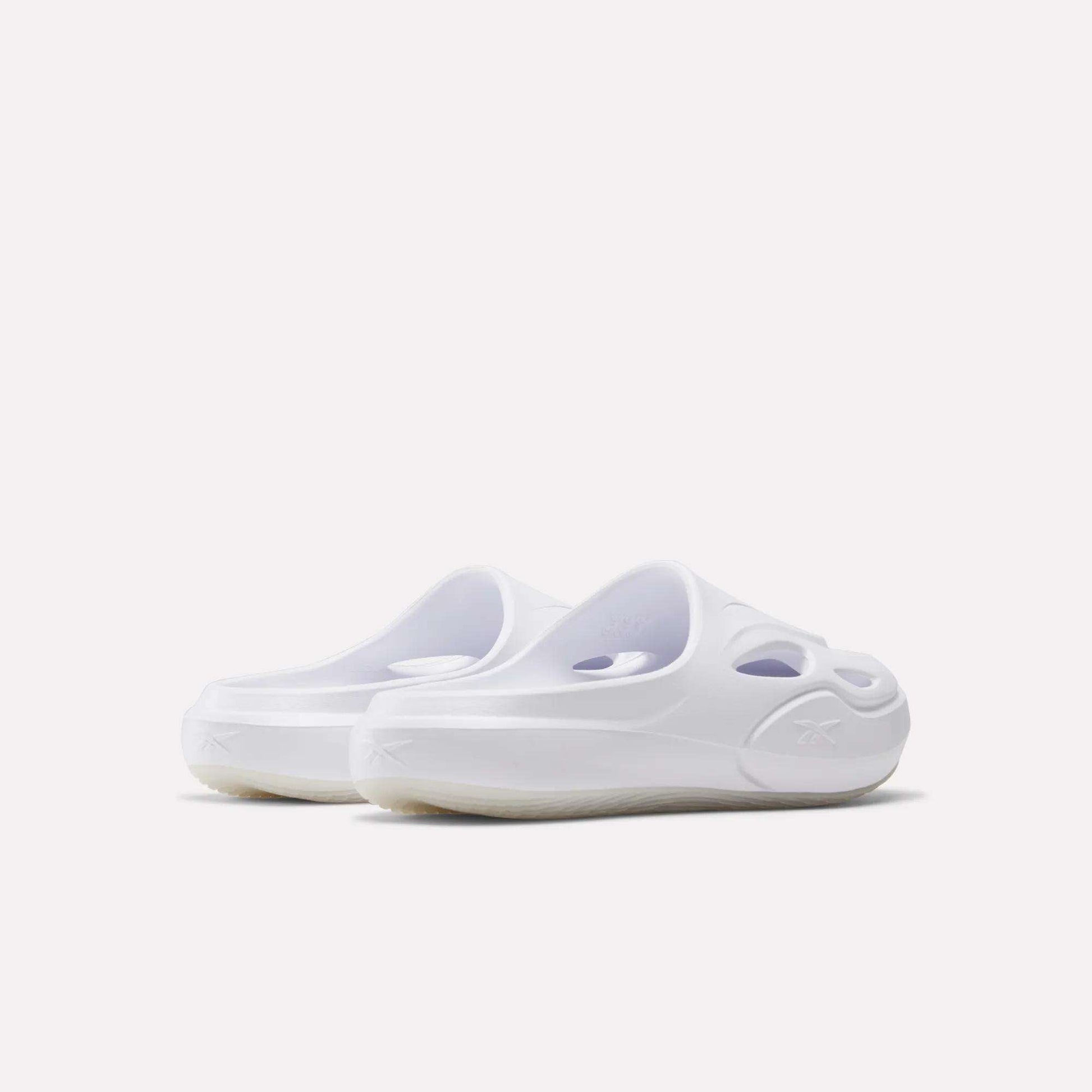 A pair of Reebok DMX Recovery Mules in white, featuring a slip-on design, molded EVA construction, cut-out details, and rounded toes is shown from an angled rear view on a plain white background.