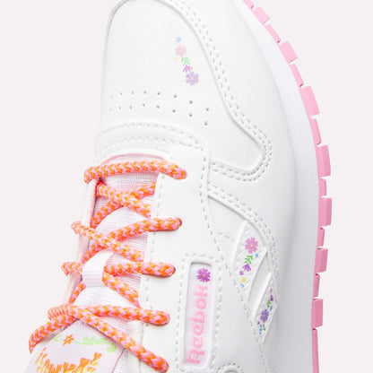 Close-up of Reebok x DORA Classic Leather Shoes for little kids, featuring white uppers, colorful floral designs, pink and orange laces, a pink-tread sole, and Reebok branding for a playful look.