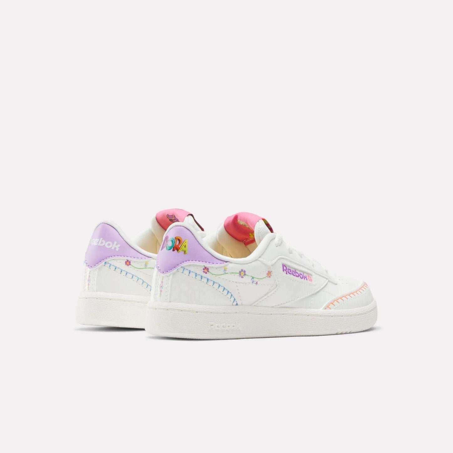The Reebok x DORA Club C Shoes for little kids feature a white base with pastel rainbow details, embroidered accents, colorful heel designs, multicolored stitching, and bold Reebok branding for a playful standout look.