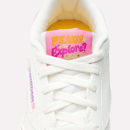 Close-up of Reebok x DORA Club C Shoes for little kids, featuring a white design with a pink and yellow tongue label reading “READY TO explore?”, playful flower and leaf accents, white laces, and a bright yellow inner lining.