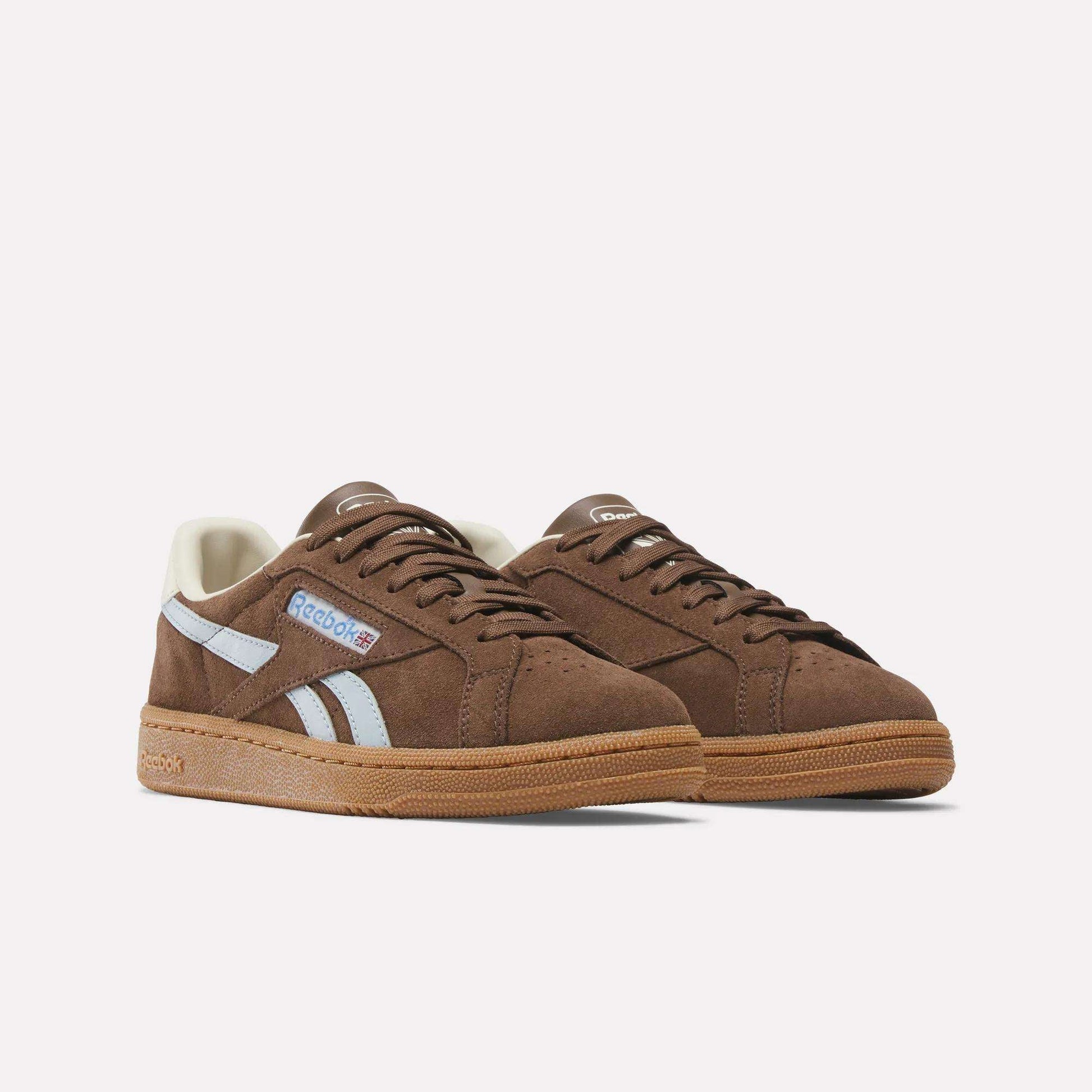 A pair of Reebok Women's Club C Grounds UK brown suede shoes with white side stripes, gum rubber soles, and Reebok logos on the side and tongue, displayed against a plain white background.