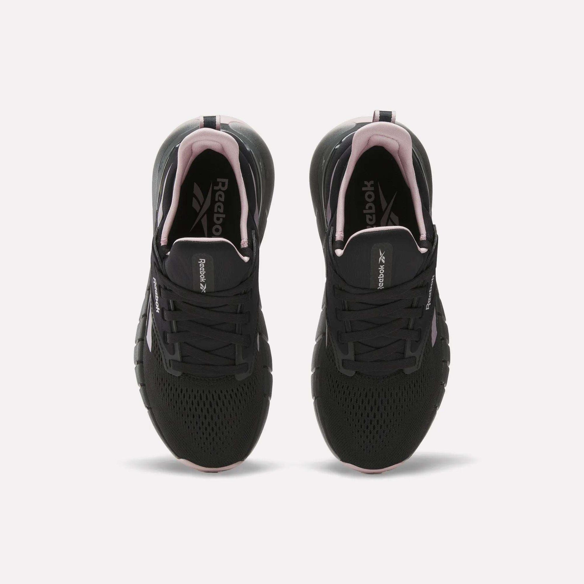 A pair of Reebok Women's Nano Gym Training Shoes in black with white soles and pink accents, shown from above on a white background. Versatile for workouts, they feature the Reebok logo on the insoles.