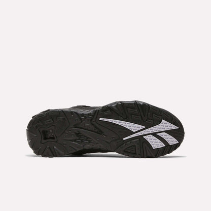 The image shows the black rubber outsole of the Reebok Brutus Low Shoes, featuring textured treads, white angular accents, and grooves for traction, blending classic heritage style with modern street-ready design.