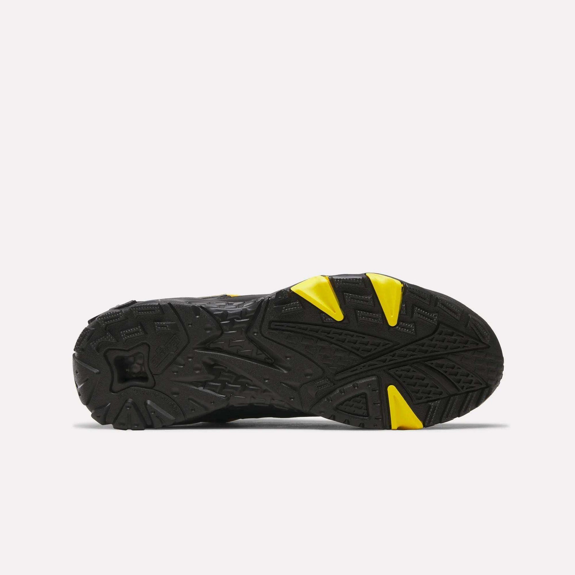 The image shows the black rubber outsole of Reebok Brutus Low Shoes, featuring distinct textured patterns and yellow accents at the toe and midfoot, highlighting its heritage design against a plain white background.
