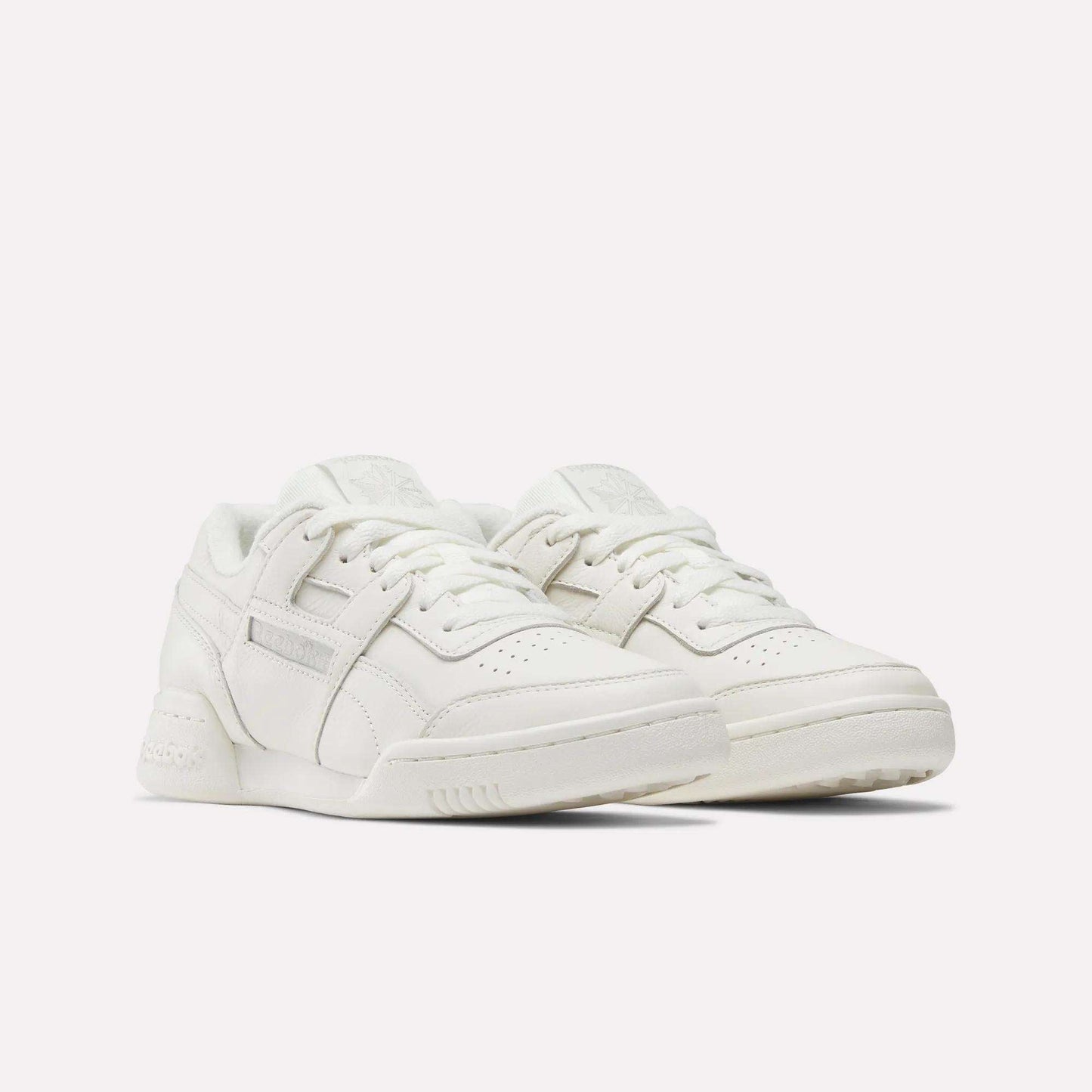 A pair of Women's Workout Plus Shoes by Reebok, featuring a white low-top design with laces, perforated toe boxes, and subtle stitching details, displayed against a plain white background.