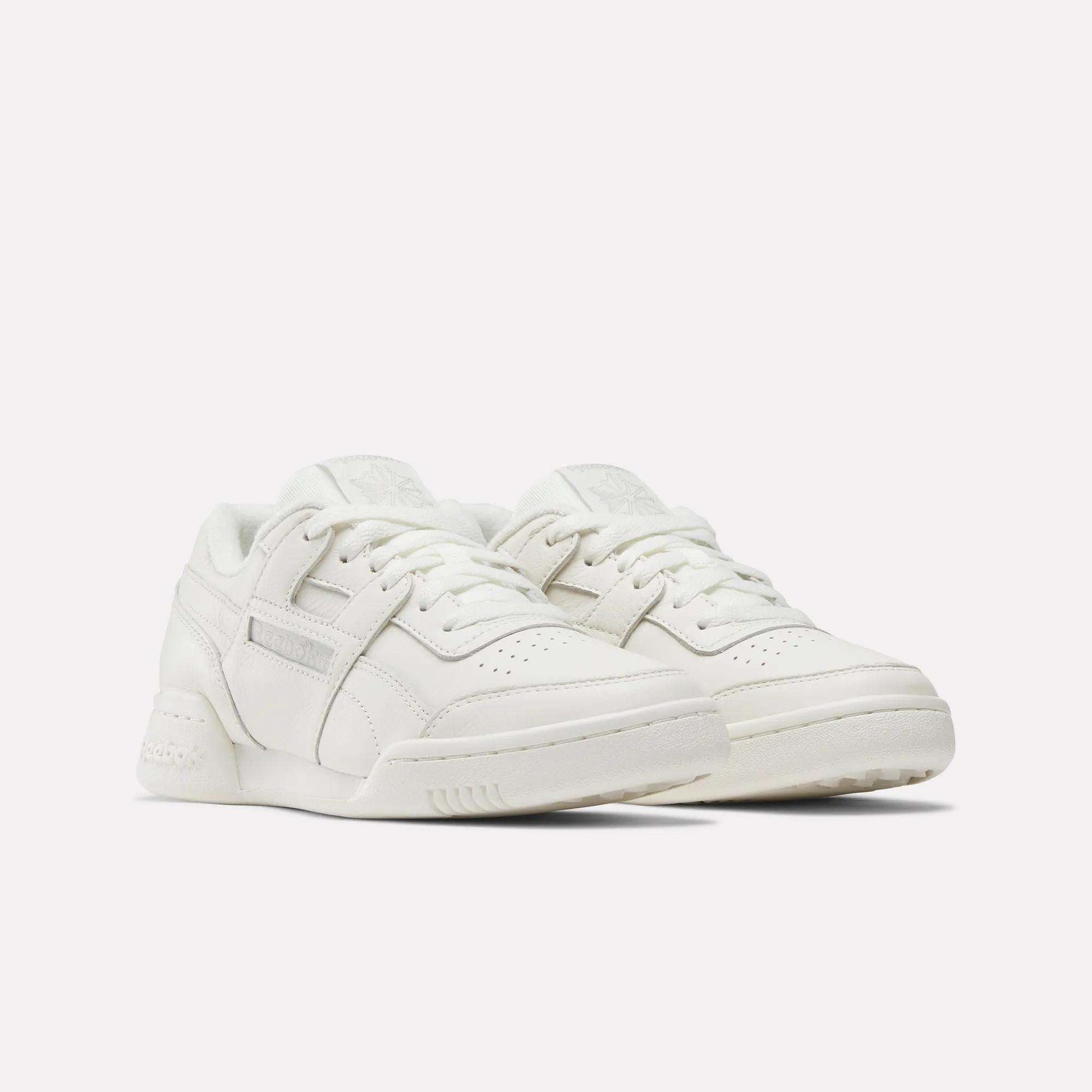 A pair of Women's Workout Plus Shoes by Reebok, featuring a white low-top design with laces, perforated toe boxes, and subtle stitching details, displayed against a plain white background.