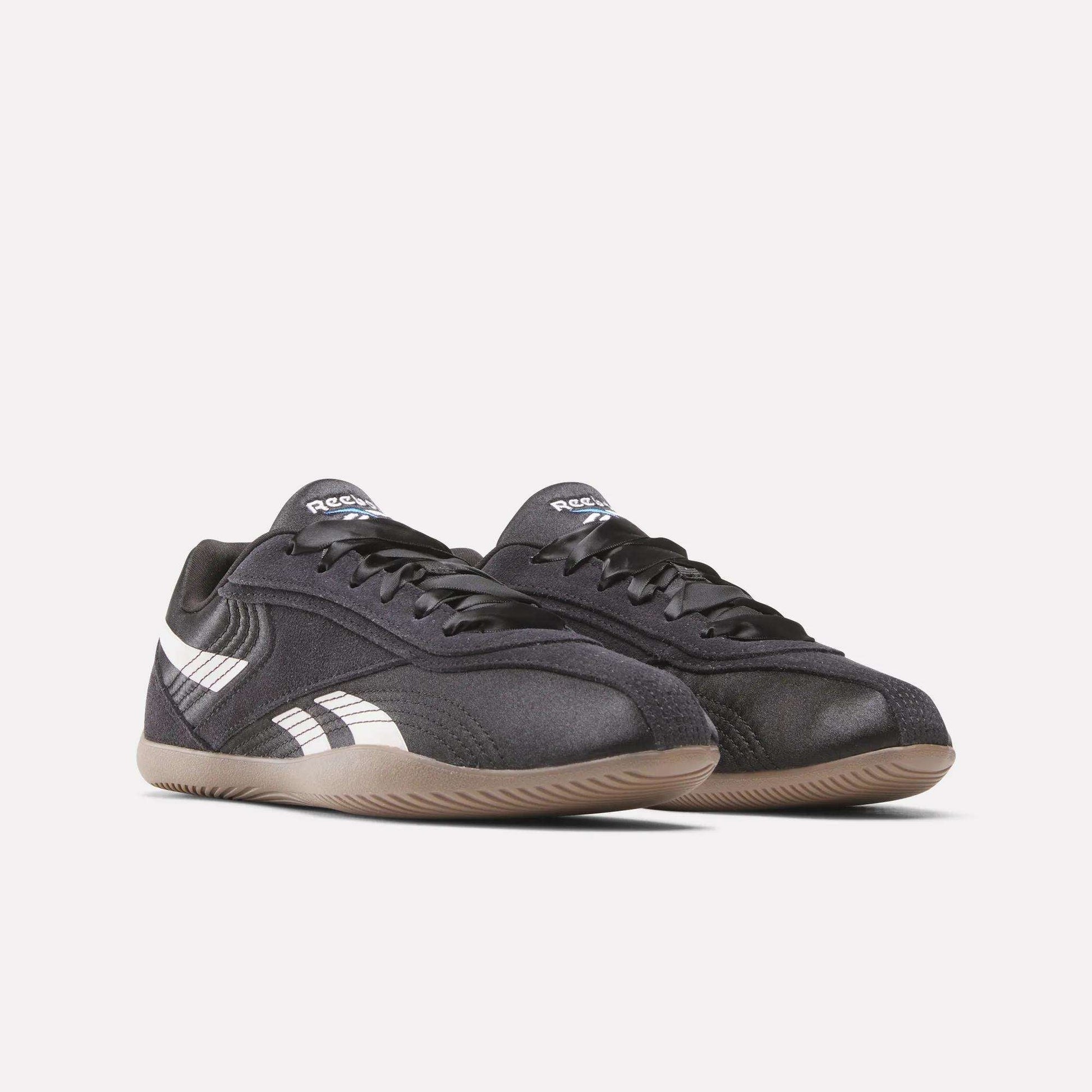 A pair of Women's Ultra Lo Shoes by Reebok in black with white side stripes, black laces, and light brown rubber soles, shown on a plain white background. Sport-inspired style for any outfit.