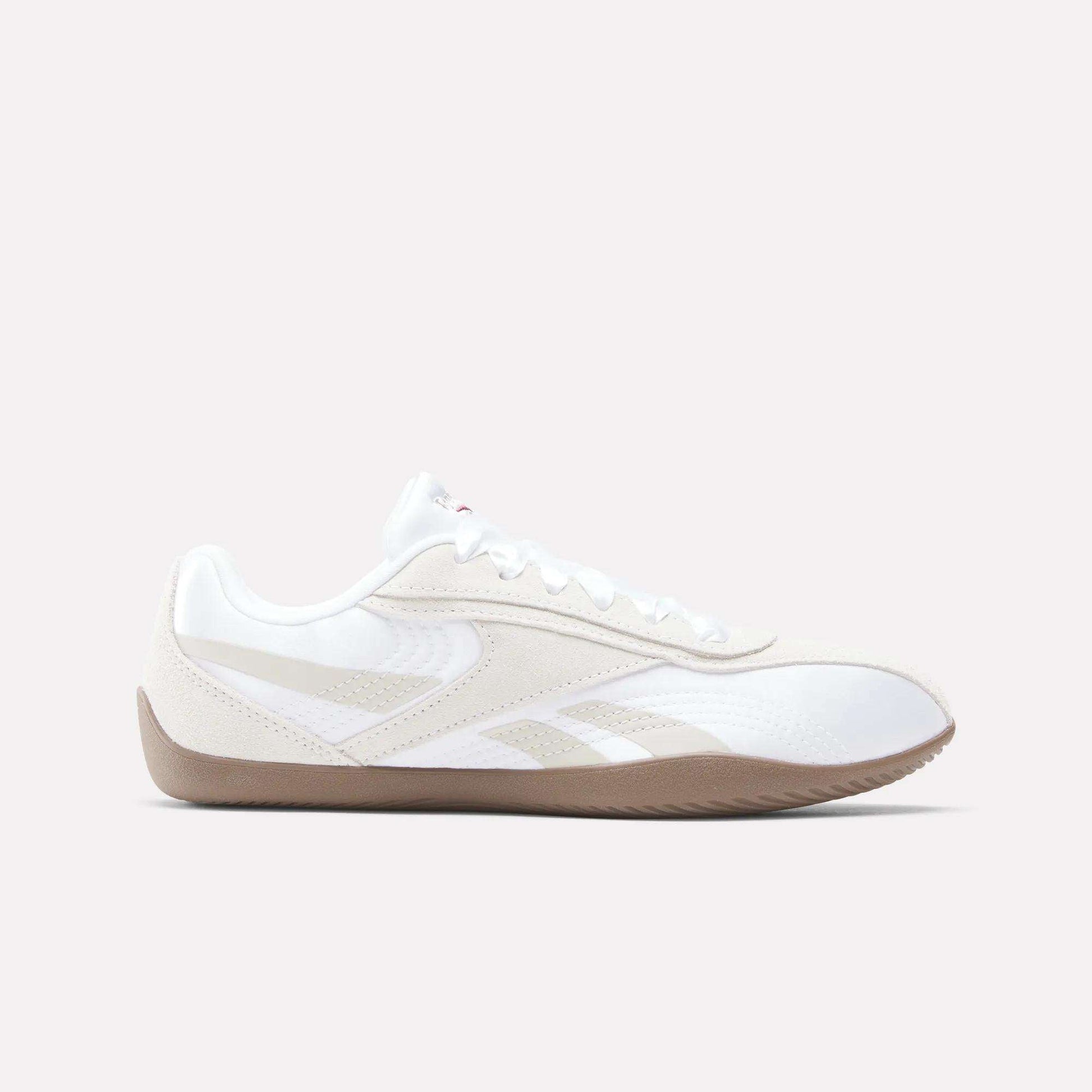 The Reebok Women's Ultra Lo Shoes feature a white athletic design with beige accents, a gum rubber sole, and lace-up closure—blending sporty style with a sleek, low-profile silhouette. Shown in side view on a light background.