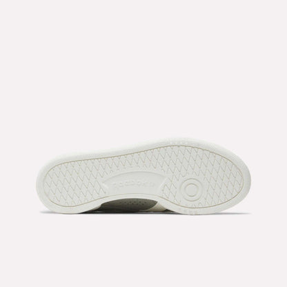 The image displays the white rubber outsole of Reebok Women's Club C Grounds UK Shoes, with a circular tread near the heel, herringbone traction patterns, and an embossed Reebok logo at the center.