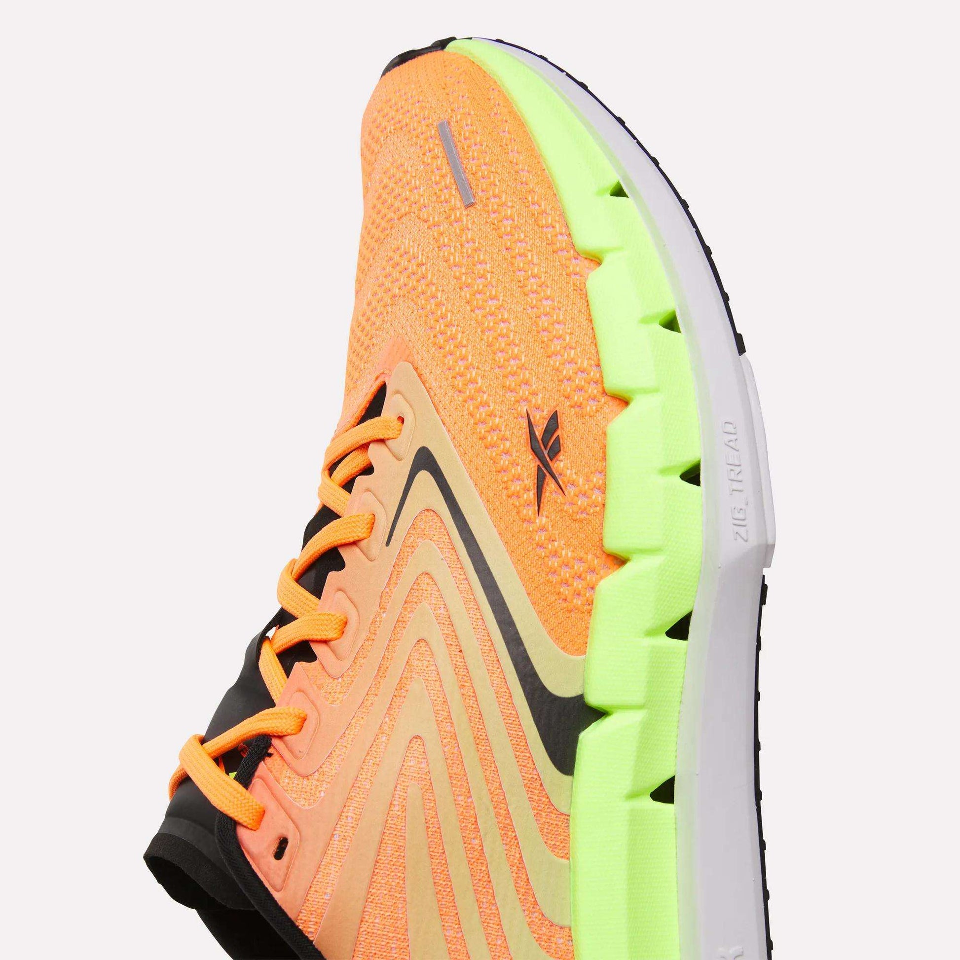 A close-up of Reebok Men's FloatZig Tread Running Shoes with an orange knit upper, black accents, and a bright green segmented sole. The cushioned midsole and wavy design offer comfort and style—ideal for treadmill workouts.