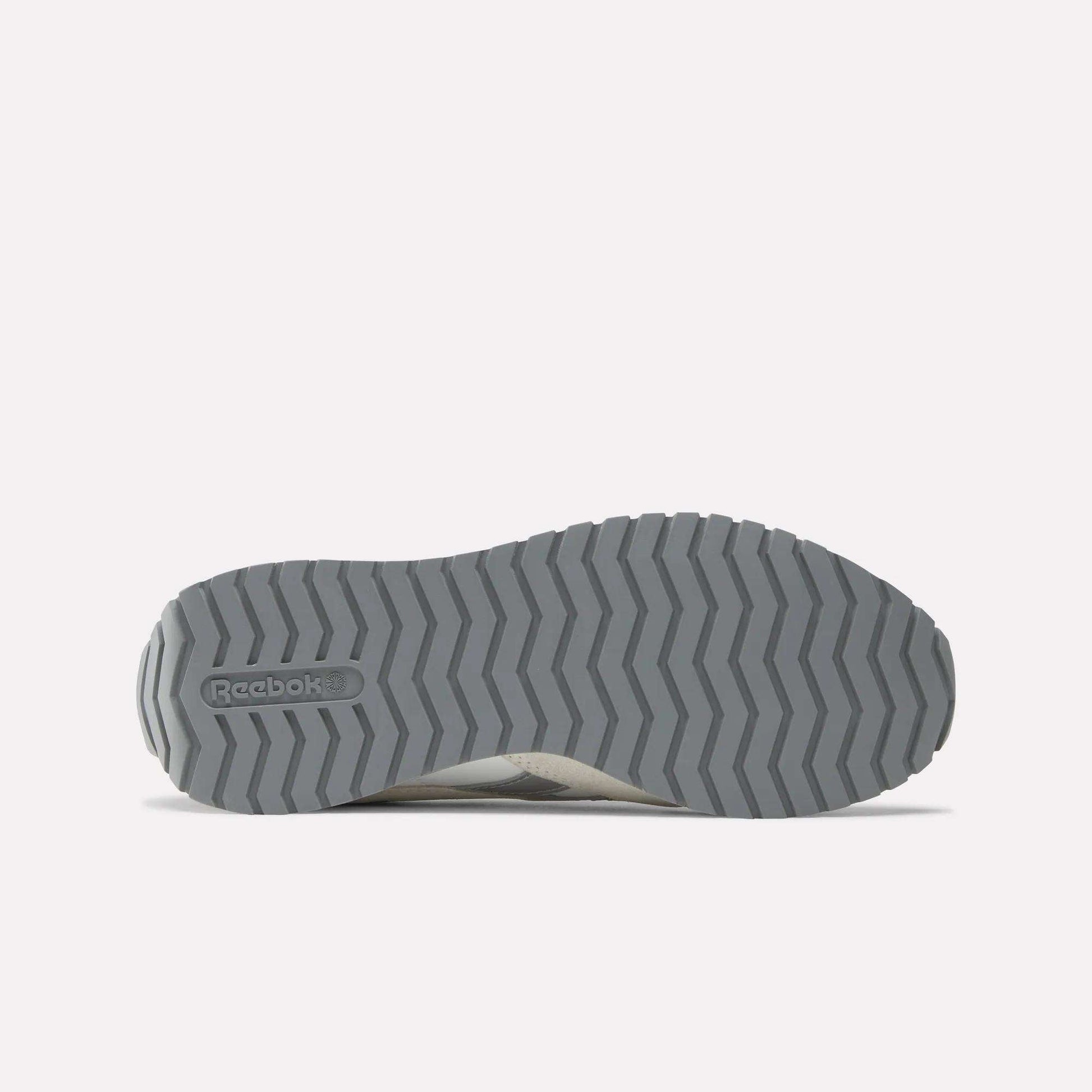 The image shows the gray rubber outsole of Reebok Women's Classic AZ Shoes with a zigzag tread and embossed logo, set against a light gray background that highlights these vintage style sneakers.